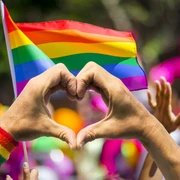 LGBTQ+ heart Hände formen ein Herz vor einer RegenbogenflaggeHands form a heart in front of a rainbow flag