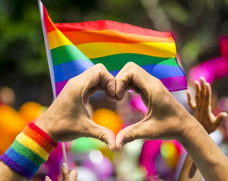 LGBTQ+ heart Hände formen ein Herz vor einer RegenbogenflaggeHands form a heart in front of a rainbow flag
