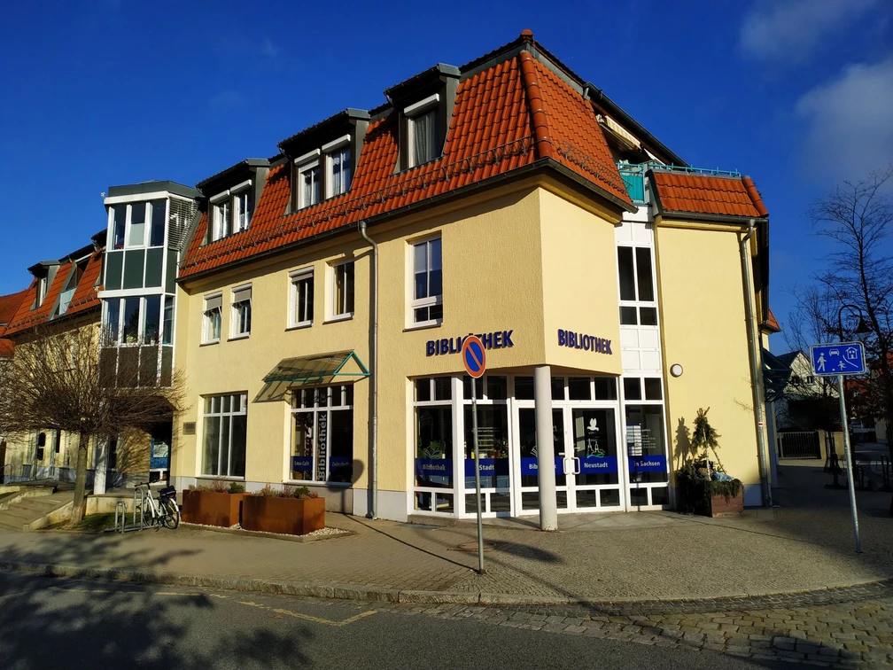 Stadtbibliothek Neustadt in Sachsen  Gelbes Gebäude mit rotem Dach, beschriftet mit "Bibliothek", bei sonnigem Wetter; Fahrradständer und Verkehrsschilder im Vordergrund.