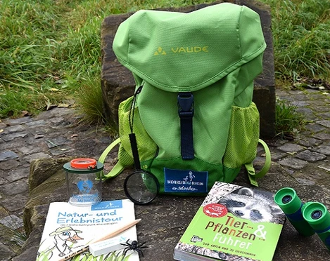 Entdeckerrucksack am Naturerlebnispfad in Monheim am Rhein Ein grüner Rucksack mit Büchern, einer Lupe und einem Fernglas liegt auf einem Naturpfad.