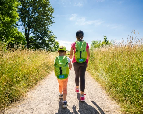 Mit dem Entdeckerrucksack auf dem Naturerlebnispfad in Monheim am Rhein Zwei Personen mit Rucksäcken spazieren auf einem sonnigen Pfad durch hohe Gräser.