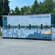 Fahrradgarage Pirna Fahrradgarage mit nummerierten Fächern und blauer Wandgestaltung, umgeben von Bäumen und gepflastertem Boden, bei klarem Himmel.