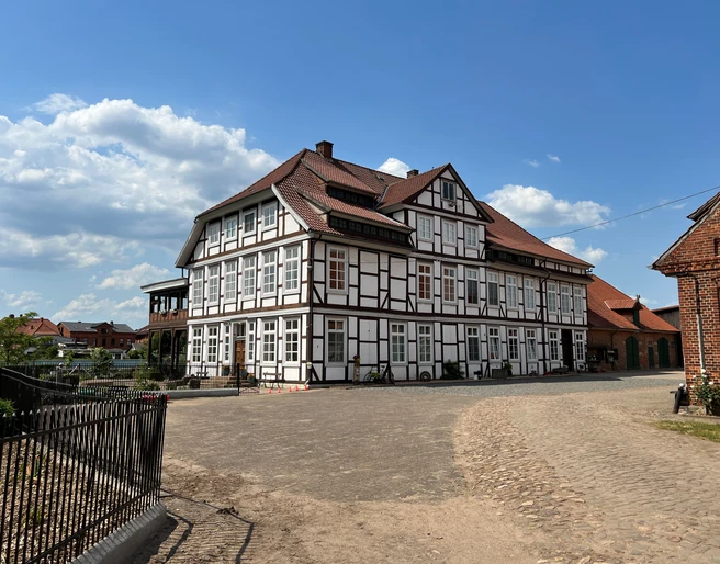 Rittergut von Behr Hoya Fachwerkhaus mit rotem Dach, umgeben von gepflastertem Hof unter blauem Himmel mit Wolken.