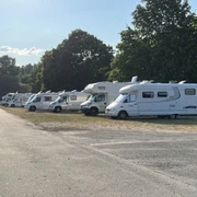 Wohnmobile stehen nebeneinander auf einer sonnigen Wiese, umgeben von Bäumen und blauem Himmel.