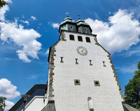 Der weiße Kirchturm der Katholischen Kirche St. Johannes Nepomuk ragt vor einem blauen Himmel empor.