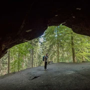 Diebskeller Diebshöhle Eine Person mit Rucksack steht am Eingang einer dunklen Höhle, umgeben von einem grünen Wald bei Tageslicht.