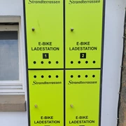 Vier leuchtend grüne E-Bike Ladesäulen der Marke Strandterrassen vor einer verputzten Wand.