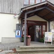Touristinformation Gohrisch Eingang einer Touristinformation mit Überdachung, Blumenkübeln und Infotafel; das Gebäude hat eine helle Fassade und Holzverkleidung.