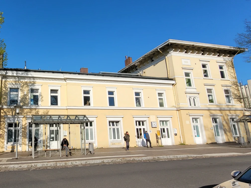 Bahnhof Ratzeburg_1980x1080_Silvana Dust_20230510 (2).jpg Leicht gelb gestrichenes Bahnhofsgebäude mit weißen Fenstern. Vor dem Gebäude ist eine Bushaltestelle