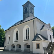 Garnisionskirche auf der Festung Königstein Weiße Kirche mit grauem Turm und Spitzdach, umgeben von Bäumen unter klarem, blauem Himmel; ruhige, sonnige Atmosphäre.