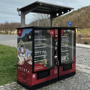 Proviantomat in Bad Schandau Roter Verkaufsautomat mit regionalen Produkten, umgeben von gepflastertem Platz und grüner Wiese, im Hintergrund Fluss und bewaldete Hügel.