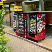 Proviantomat am Parkhaus der Festung Königstein Zwei rote Verkaufsautomaten mit Snacks und Getränken stehen neben einem Geldautomaten vor einem Gebäude, umgeben von grüner Vegetation.