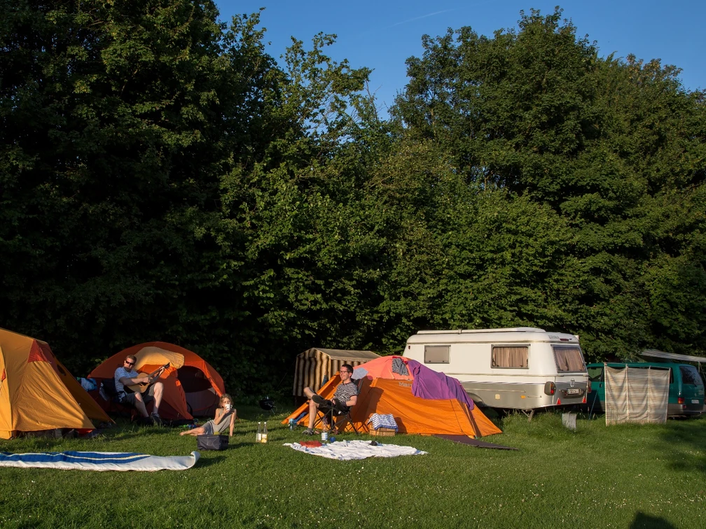 Camping 3 ©Jens Butz.jpg Auf dem Campingplatz stehen mehrere Zelte und ein Wohnanhänger, umgeben von Personen, die sich im Freien aufhalten