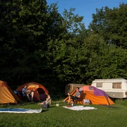 Camping 3 ©Jens Butz.jpg Auf dem Campingplatz stehen mehrere Zelte und ein Wohnanhänger, umgeben von Personen, die sich im Freien aufhalten