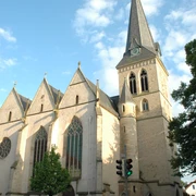 Historische Kirche mit gotischen Elementen, markantem Turm und grünen Bäumen unter blauem Himmel.
