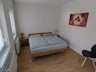 Burkis Ferienhaus Ein helles Schlafzimmer mit Doppelbett, zwei Nachttischen, Stuhl und Kunstbild an der Wand.