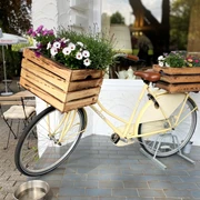 Restaurant Wohnzimmer Neviges Ein vintage gelbes Fahrrad, dekoriert mit blühenden Holzblumenkästen, lehnt an einer weißen Wand.