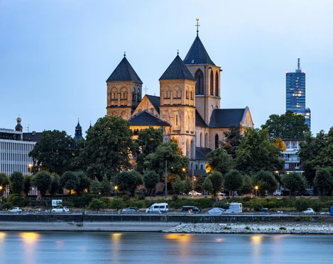 Saint Kunibert Die romanische Kirche Sankt Kunibert in Köln erhebt sich mit ihren markanten Türmen über den Rhein, flankiert von moderner Architektur im Hintergrund.The Romanesque church of St. Kunibert in Cologne rises above the Rhine with its striking towers, flanked by modern architecture in the background.