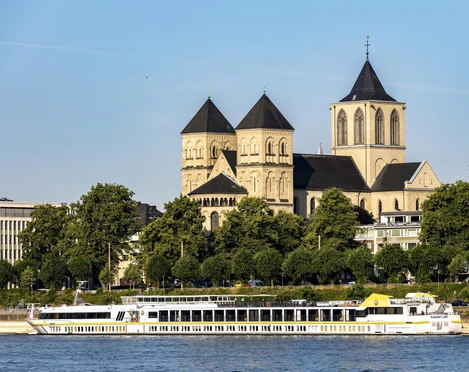Saint Kunibert Ein romanisches Kirchenbauwerk mit drei Türmen hebt sich majestätisch vor einem klaren Himmel ab. Davor gleitet ein elegantes Ausflugsboot sanft entlang des Rheins, während grüne Bäume das Ufer säumen.A Romanesque church with three towers stands out majestically against a clear sky. In front of it, an elegant excursion boat glides gently along the Rhine, while green trees line the banks.