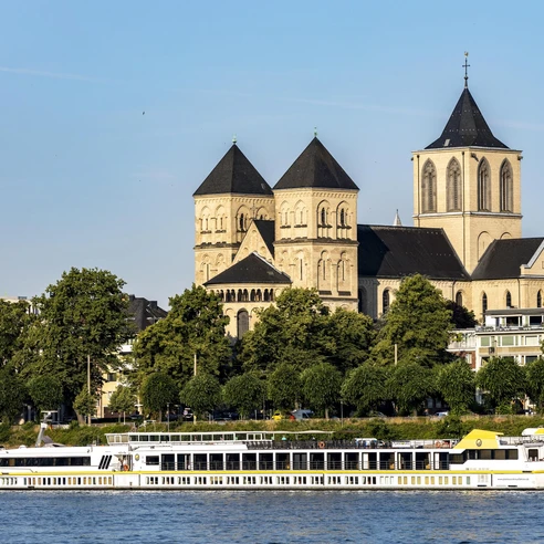 Saint Kunibert Ein romanisches Kirchenbauwerk mit drei Türmen hebt sich majestätisch vor einem klaren Himmel ab. Davor gleitet ein elegantes Ausflugsboot sanft entlang des Rheins, während grüne Bäume das Ufer säumen.A Romanesque church with three towers stands out majestically against a clear sky. In front of it, an elegant excursion boat glides gently along the Rhine, while green trees line the banks.