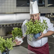DSCF6525 Küche final.jpg Ein Koch in Uniform präsentiert stolz frische Kräutertöpfe in einer modernen Edelstahlküche.