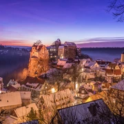 Ausblick auf Hohnstein im Winter Verschneite Stadt bei Sonnenuntergang mit beleuchteten Fachwerkhäusern, einer Kirche und einer Burg auf einem Hügel, umgeben von Wald.