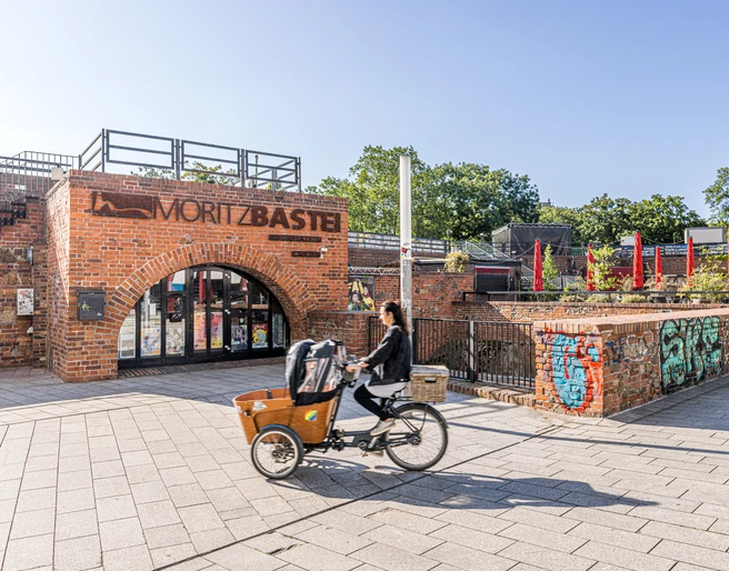 Moritzbastei - Veranstaltungen in Leipzig Bild vom oberen Eingang zur Moritzbastei am Kurt-Masur-Platz in Leipzig, in der es ein Café und Restaurant gibt und in dem viele Veranstaltungen stattfinden, eine Fahrradfahrerin fährt durch das Bild