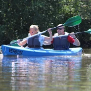 Kanutour Ein Mann und eine Frau paddeln in einem blauen Kanu durch ein ruhiges Gewässer, umgeben von grüner Natur.