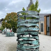 Erinneringe in Mettmann Eine skulpturale Bronze-Säule mit gravierten Jahreszahlen steht vor einer Kirche bei bewölktem Himmel.