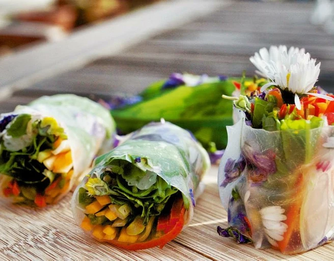 Kräuter-Tapas Das Bild zeigt drei frische Frühlingsrollen mit buntem Gemüse und essbaren Blüten auf einem Holztisch.
