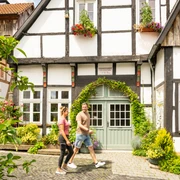 Tecklenburg-Altstadt-Tecklenburger Land, D. Ketz-2023-120.jpg Fachwerkhäuser mit Blumen und Grünpflanzen dekoriert, ein Paar spaziert eine gepflasterte Gasse entlang.