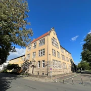 Dreistöckiges historisches Schulgebäude mit gelber Fassade und markantem Giebel unter blauem Himmel.
