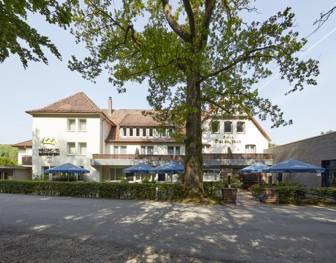 21008_Waldhotel_Baerenstein_Aussen_02.jpg Das Waldhotel Bärenstein mit umgebenden Bäumen und Terrasse mit Sonnenschirmen bei Tageslicht.