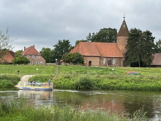 Kirche Westen Backsteinkirche mit Spitzdach und Turm an Flussufer, kleines Boot am Steg, umgeben von Grünflächen.