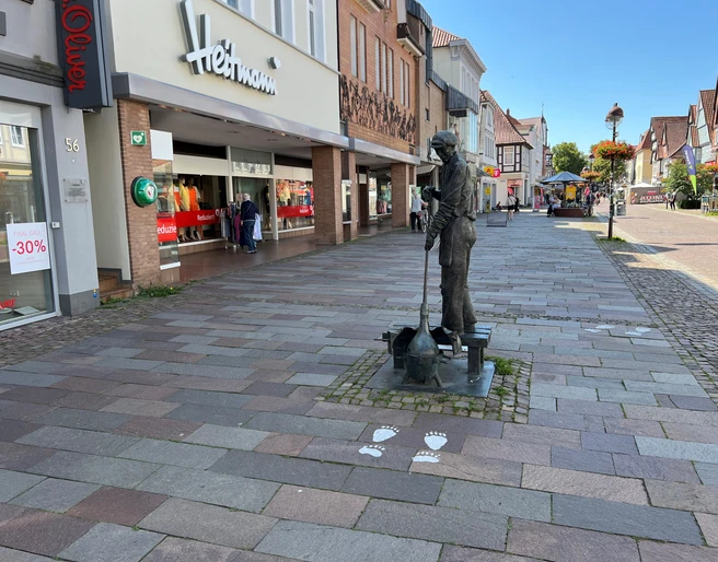Glasbläser Eine Bronzestatue eines Glasbläsers steht auf einem steinernen Gehweg in einer Einkaufsstraße.