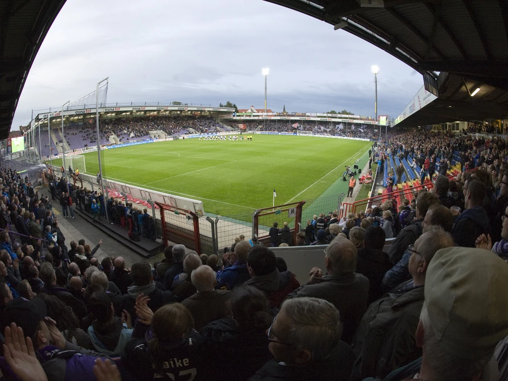 Fußballstadion bei Abendlicht mit vollen Tribünen und beleuchtetem Spielfeld, Fans jubeln lebhaft.