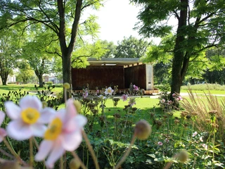 Der SolePavillon im Kurpark in Bad Laer Rostfarbene Stahlwand in einem sonnigen Park, umgeben von blühenden Blumen und Bäumen.