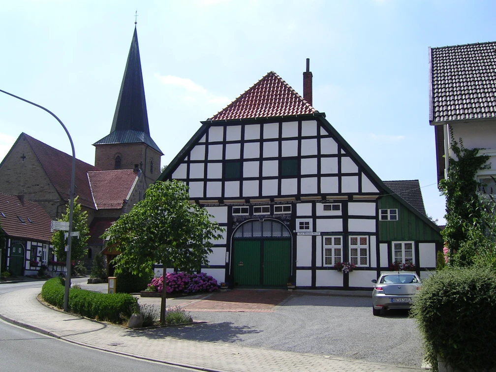 Fachwerkhaus mit grünen Akzenten, dahinter Kirchturm, am Straßenrand mit Blumen und geparktem Auto.