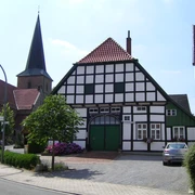 Gildehaus mit Kirche in Bissendorf Fachwerkhaus mit grünen Akzenten, dahinter Kirchturm, am Straßenrand mit Blumen und geparktem Auto.