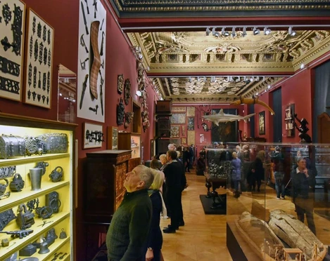 Besuchende in einem dunkelrot gestrichenen Museumsraum bewundern verschiedene Exponate, wie Krüge und Waffen, und Deckenverzierungen.