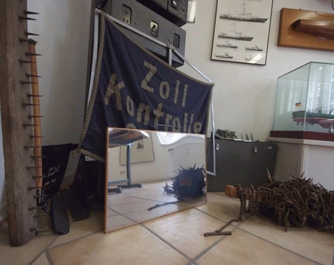Zollmuseum Friedrichs In der Ecke eines Raumes stehen verschiedene Ausstellungsstücke wie ein Schiffsmodell und ein Banner mit der Aufschrift "ZOLLKONTROLLE".