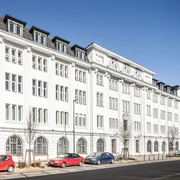 Stadtarchiv Aachen Helles, langes, historisches Gebäude mit vier Stockwerken und hohen Fenstern mit Sprossen. Vor dem Gebäude stehen einige parkende Autos.