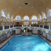 Historisches Hallenbad mit hohem Gewölbe, Arkadenfenstern und zentralem Schwimmbecken.