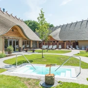 Außenbereich eines ländlichen Wellnesshotels mit Reetdach, Pool und Liegestühlen.