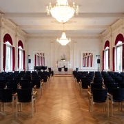 spiegelsaal-bestuhlt Der elegant bestuhlte Spiegelsaal, geprägt vom leuchtenden Kronleuchter und großen Fenstern mit roten Vorhängen.