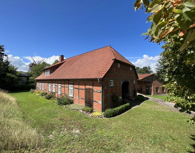 Historisches Backsteinhaus mit rotem Satteldach, umgeben von grüner Wiese, unter blauem Himmel.