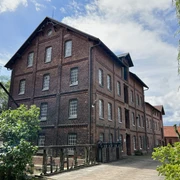Wassermühle Stemmen Alte, dreistöckige Backsteinmühle mit Satteldach, umgeben von Bäumen und einem Holzbalkon im Vordergrund.
