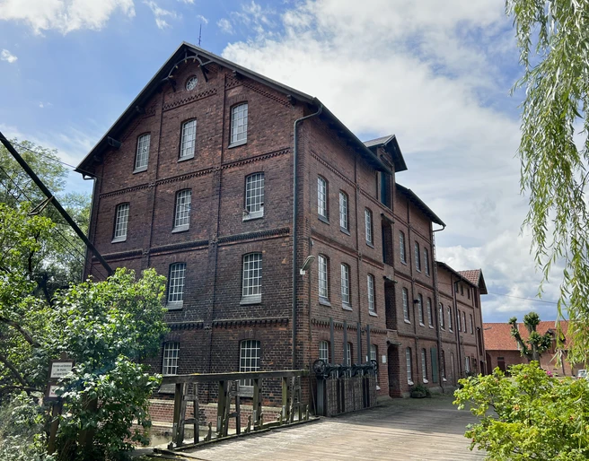Wassermühle Stemmen Alte, dreistöckige Backsteinmühle mit Satteldach, umgeben von Bäumen und einem Holzbalkon im Vordergrund.
