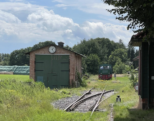 Ein alter Güterschuppen mit grünen Toren und Uhr, daneben ein Schienenweg mit einer historischen Lokomotive.