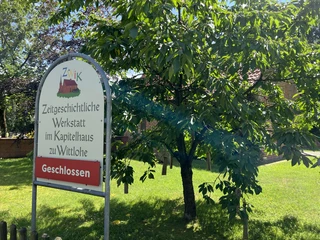 Kapitelhaus Wittlohe Schild vor einem historischen Gebäude in Wittlohe, umgeben von grünen Bäumen und Sonnenschein.